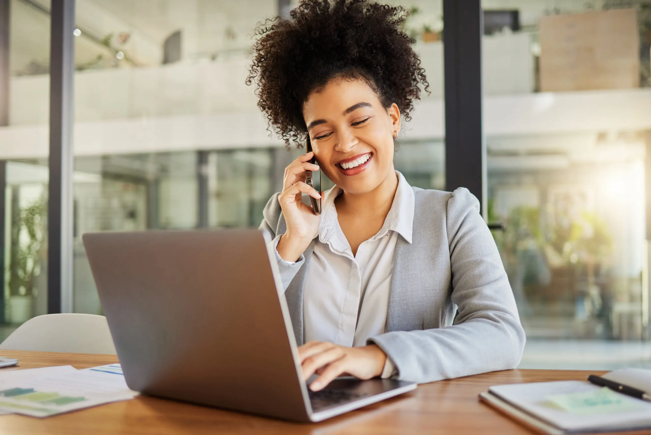 Business woman working on laptop in office, in conversation on phone and talking with people on smartphone at work. Happy corporate worker, employee and manager reading email at marketing company A woman sitting at a table with a laptop and phone.